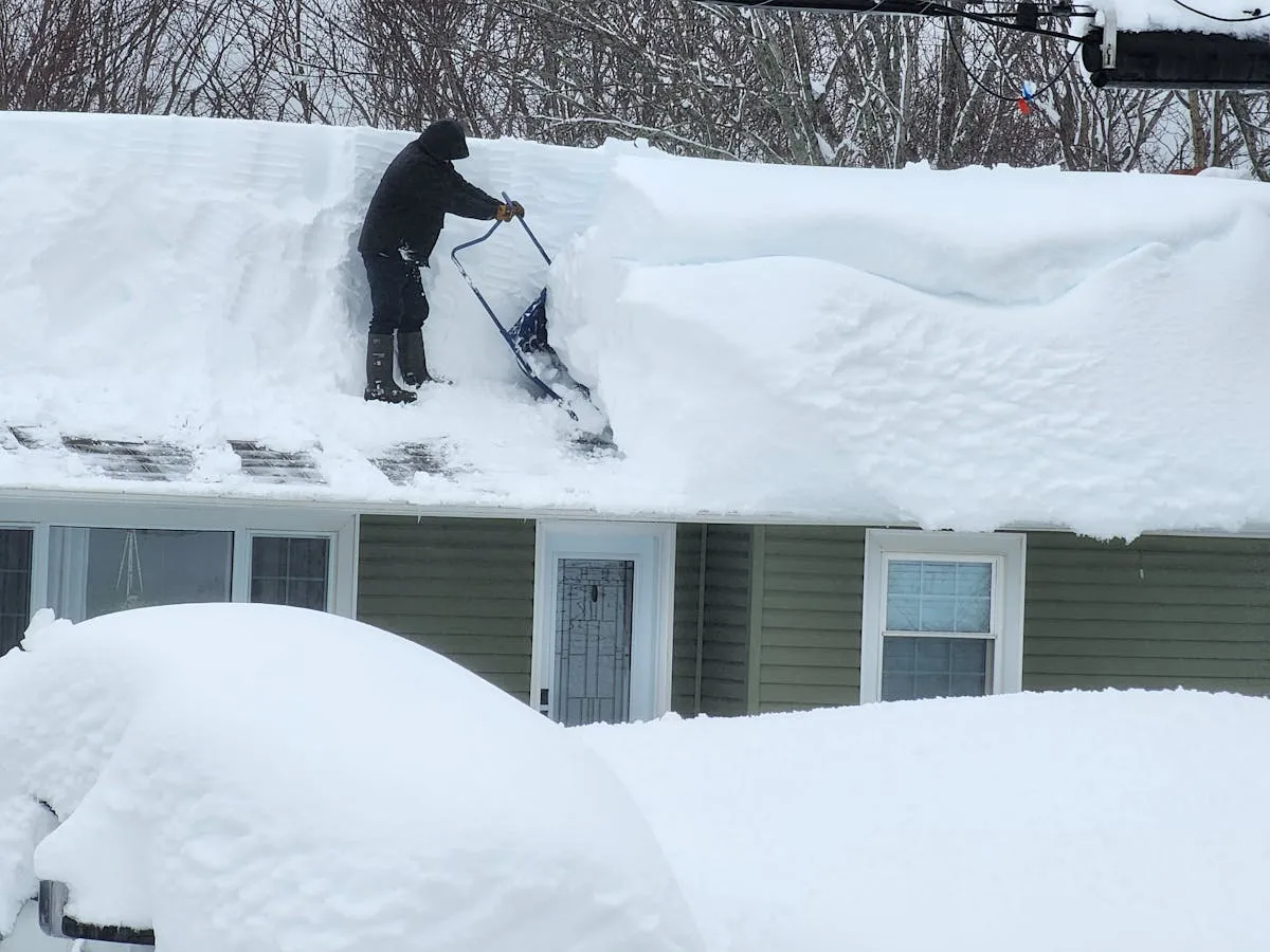 Utah Ice Dam Removal | Roof Gutter Now 13 image shows a person in dark winter clothing performing snow removal on a residential roof. The worker is using a in Utah